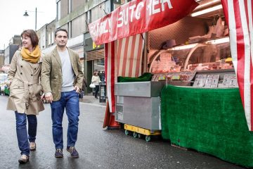 Woman wearing beige trench coat and man wearing beige jacket walking on street in London next to food cart.