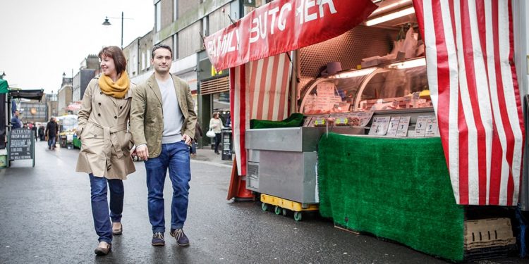 Woman wearing beige trench coat and man wearing beige jacket walking on street in London next to food cart.
