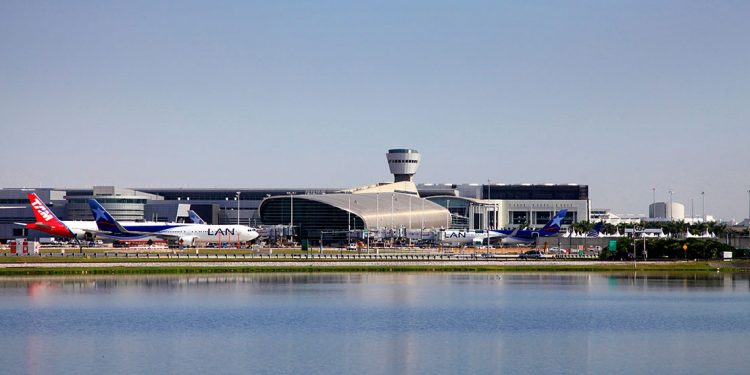 Water in foreground with three planes and silver building of airport in background.