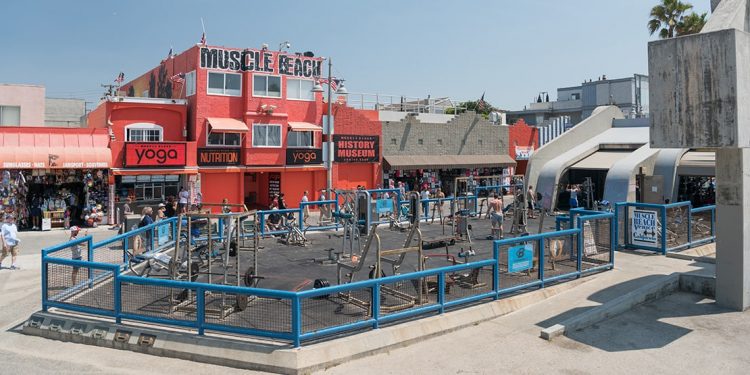 Red building with Muscle Beach painted on top of it. Blue fenced off area with fitness equipment.