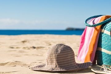 Sun hat, beach bag with towel and sunglasses hanging off of it, and sandals on the beach.