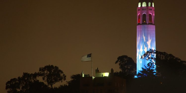 American flag blowing atop a hill with stars projected on a tower.