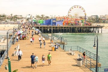 Wooden pier extending over the water with metal streetlights and wooden benches. People milling about. Beach off to the left and Ferris wheel off to the right.
