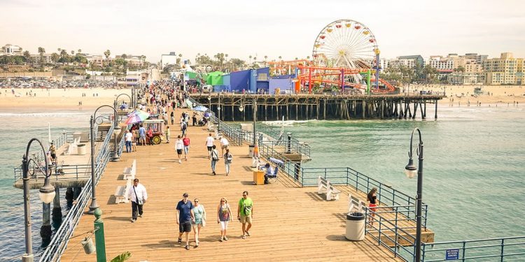 Wooden pier extending over the water with metal streetlights and wooden benches. People milling about. Beach off to the left and Ferris wheel off to the right.