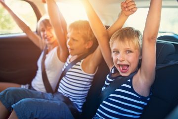 Three kids sit in the back seat of car with arms raised in excitement.