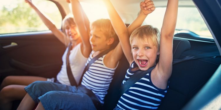 Three kids sit in the back seat of car with arms raised in excitement.