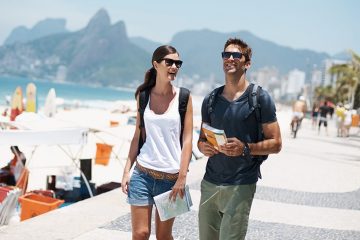 Woman and man walk along a pavement sidewalk beside the beach. She is holding a map and he is holding a book.