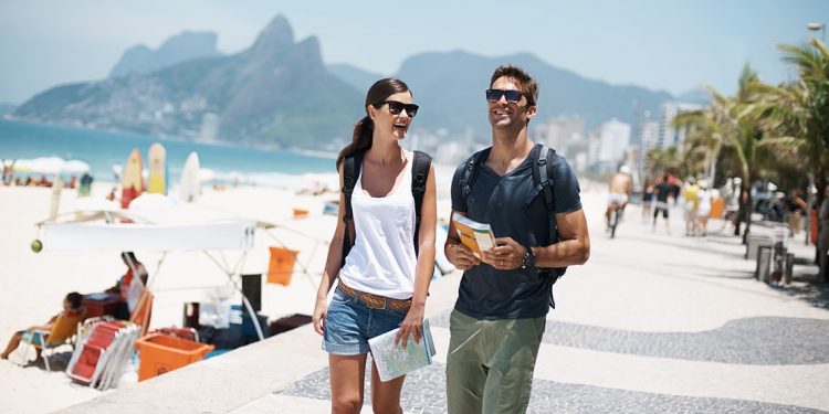 Woman and man walk along a pavement sidewalk beside the beach. She is holding a map and he is holding a book.