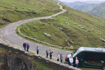Group of people standing on side of road with a van. The road winds upward through a green moor landscape.