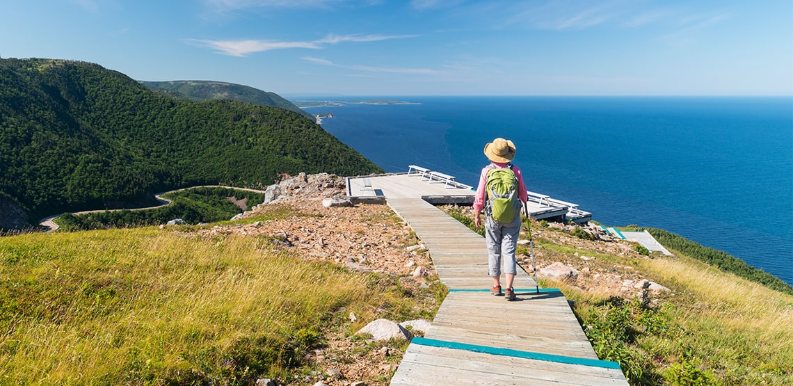 Woman with walking stick walks on boardwalk trail along crest of a hill beside the ocean. The trail continues down the hill and along the coast.
