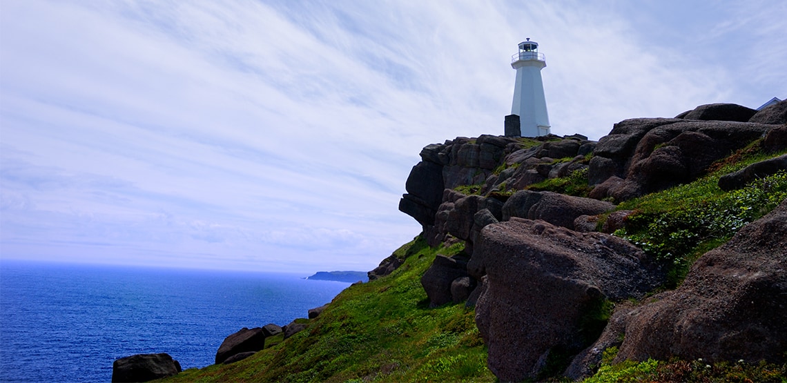 White lighthouse on a rocky outcrop atop a green hill on the coast.