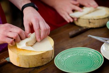 Two sets of hands rolling a spring roll on a wooden block with green plates in front of them.