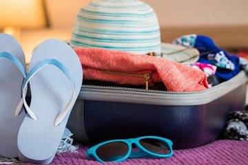 Open suitcase on a bed with sweater, swimsuits, sunhat, flip flops, and sun glasses.