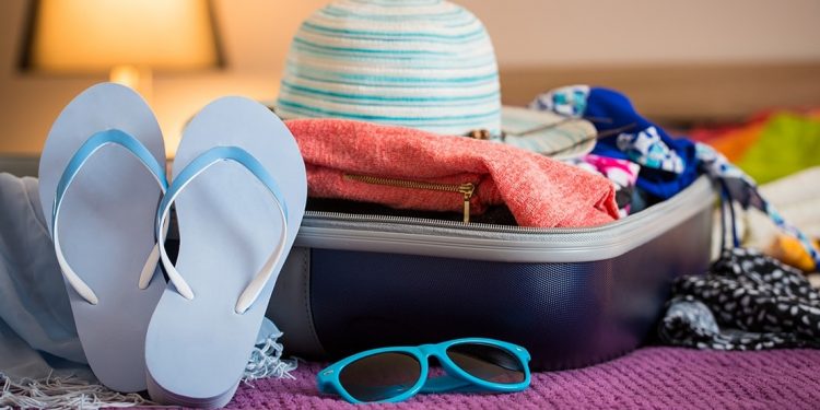 Open suitcase on a bed with sweater, swimsuits, sunhat, flip flops, and sun glasses.