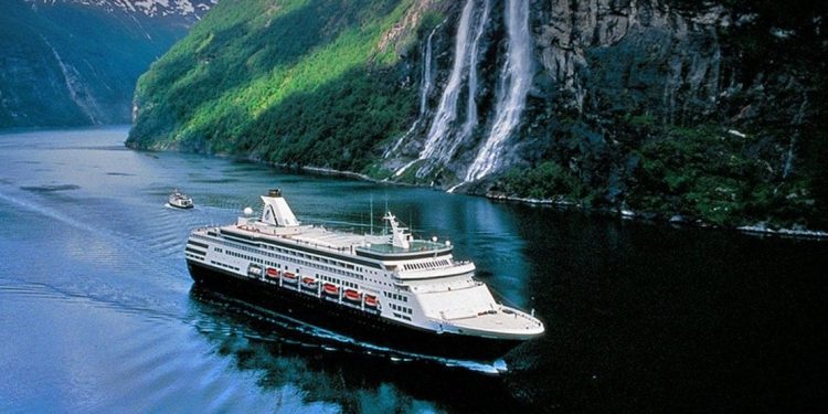 White and black cruise ship sails along a cliff with waterfalls pouring down into the water.
