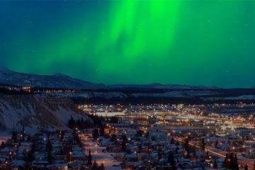 The green northern lights play over glowing lights in a city in a valley.