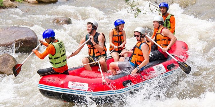 Six rafters wearing orange PFDs paddle in a red raft through raging waters.