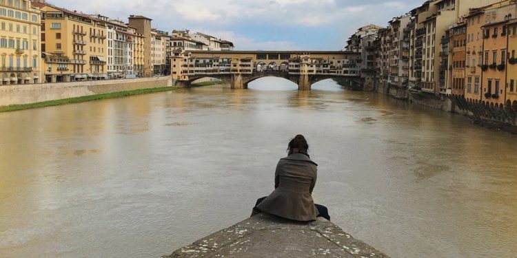 A woman sits on the edge of a piece of concrete in the middle of a river. There are joined buildings on either side and a bridge up ahead.