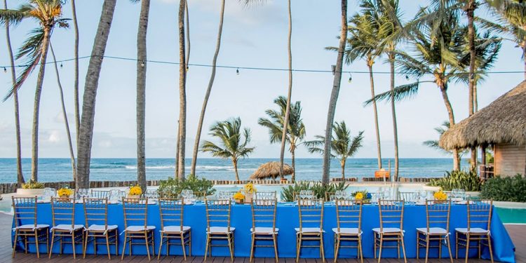 Long table with blue table cloth and chairs by the water with palm trees.