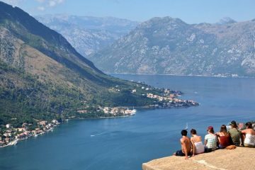 Friends sit on the edge of a rock overlooking a harbor. A mountain is on the opposite side of the water.