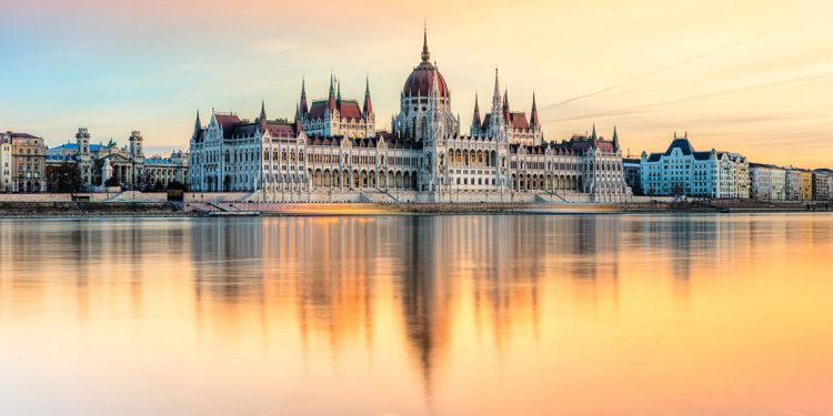 A palace with white walls and red spires on the edge of a river, the water glowing in the sunset.