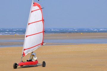 A person rides in a sailboat with wheels across the sand of a beach.