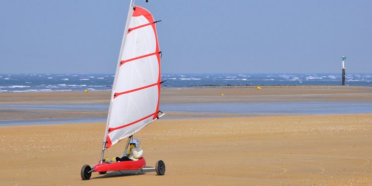 A person rides in a sailboat with wheels across the sand of a beach.