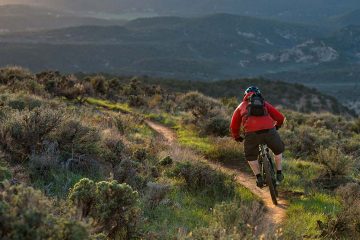 A man wearing a red jacket mountain bikes down a single track trail in a hilly landscape.