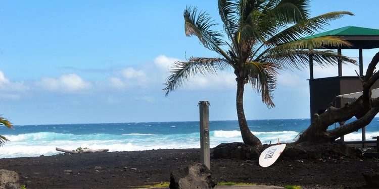 Black sand next to a crashing surf. A surfboard sits near a lifeguard hut.