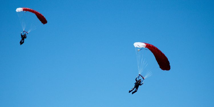 Two sky divers with red and white parachutes open against a clear blue sky.