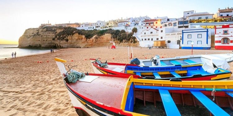 Colorful dingy boats on a beach with white washed houses with colorful trim in the background.