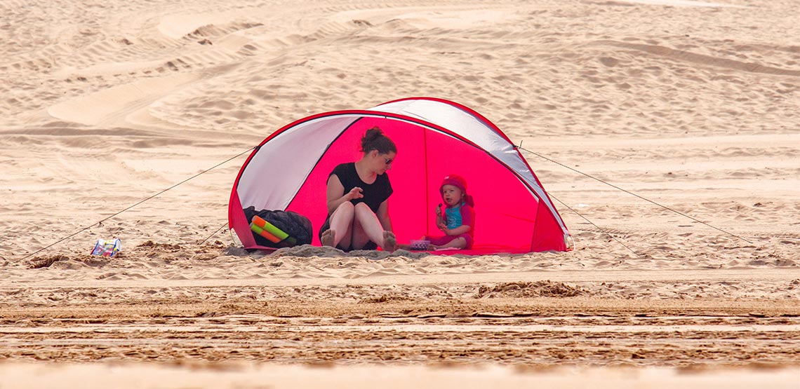 Woman and child sitting under sunshade tent on the beach.