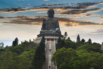 Big Buddha is a massive statue of Buddha sitting atop a mountain covered in trees with a staircase leading up to its base.