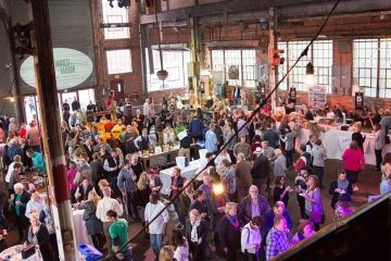 Crowds of people in a warehouse-like room where sampling tables have been set up.
