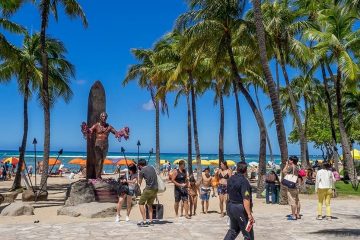 Tourists walk on a path beside Waikiki Beach and palm trees, some taking photos of a statue of a surfer.