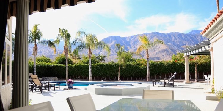 A patio in Palm Springs with a pool, hot tub and palm trees. A hedge lines the back of the pool area and mountains are visible over top.