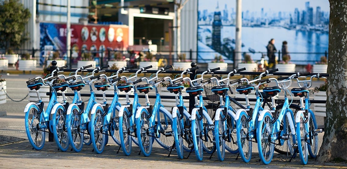 Row of blue rental bikes are lined up at the side of a road.