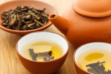 A copper colored tea pot, two bowls of steeping tea, and a bowl of loose leaf.