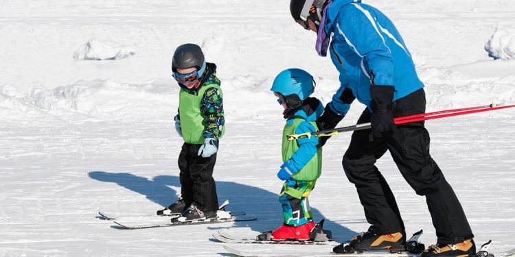 A ski instructor helping a long the youngest of two kids on skis.