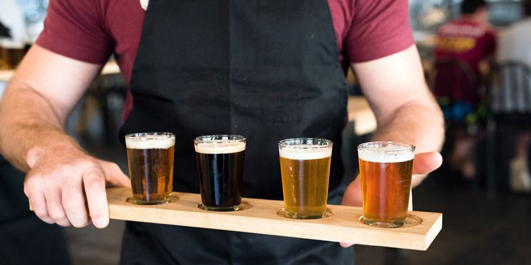 Man with black apron holding a wooden board with four small glasses filled with beer.