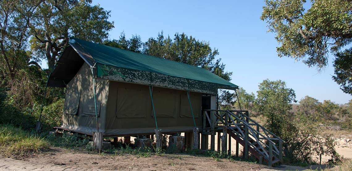 Small brown hut with green tarp roof on a wooden platform with wooden stairs leading up to it.