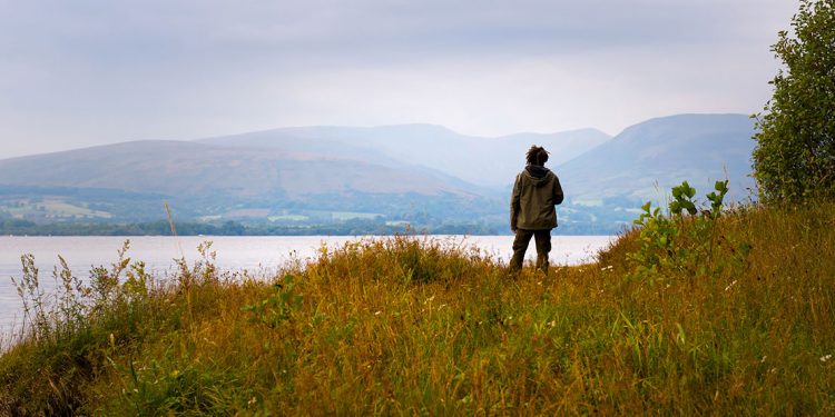 Man standing in long grass on the edge of a lake with patchwork fields on the far side and mountains rising up into hazy skies.