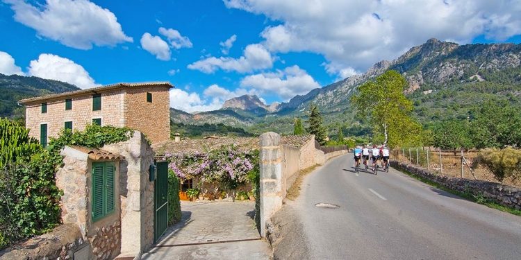 Cyclists riding along road toward mountains with Spanish style building to their left.