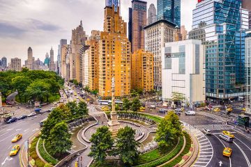 Roundabout with skyscrapers on right side and Central Park on the left. There is a ring of trees in the middle of the roundabout with a statue in the middle.