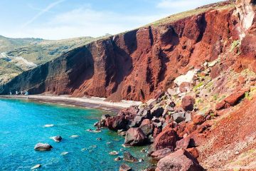 Red cliff stretching along coast of blue ocean with a strip of beach at the bottom.