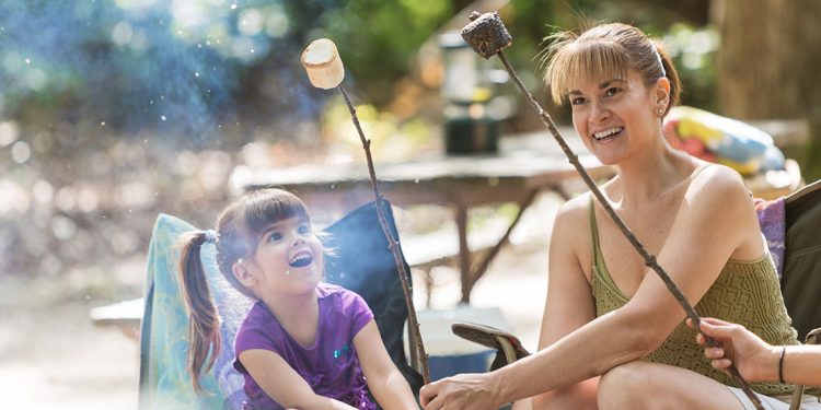 Little girl and mother sitting in lawn chairs holding sticks with roasted marshmallows on the ends.
