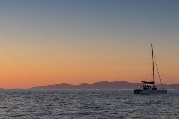 Sailboat on the ocean with silhouette of mountains in background and a peachy skyline.