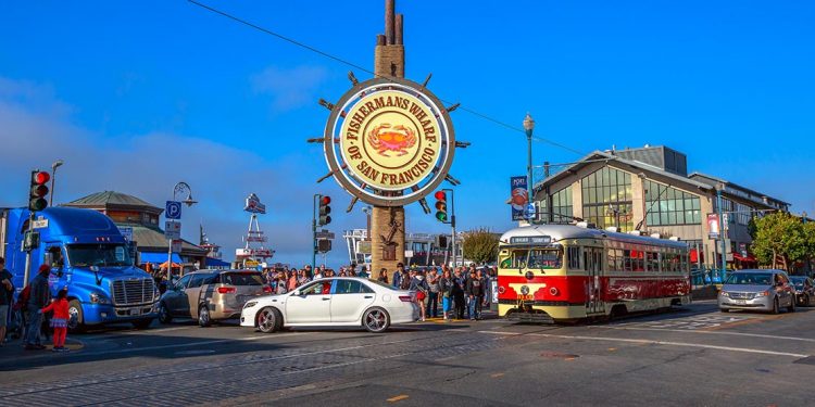 Corner of street with streetcar and giant sign reading "Fisherman's Wharf of San Francisco."