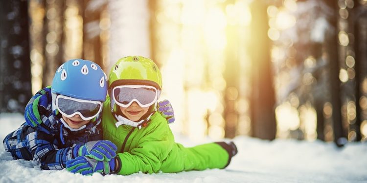 Portrait of little brothers in ski outfits lying on snow and smiling into the camera. Kids are wearing colorful helmets and ski goggles.