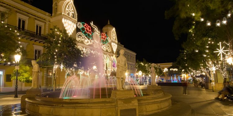 Fountain with colored lights with surrounding buildings and trees covered in lights.